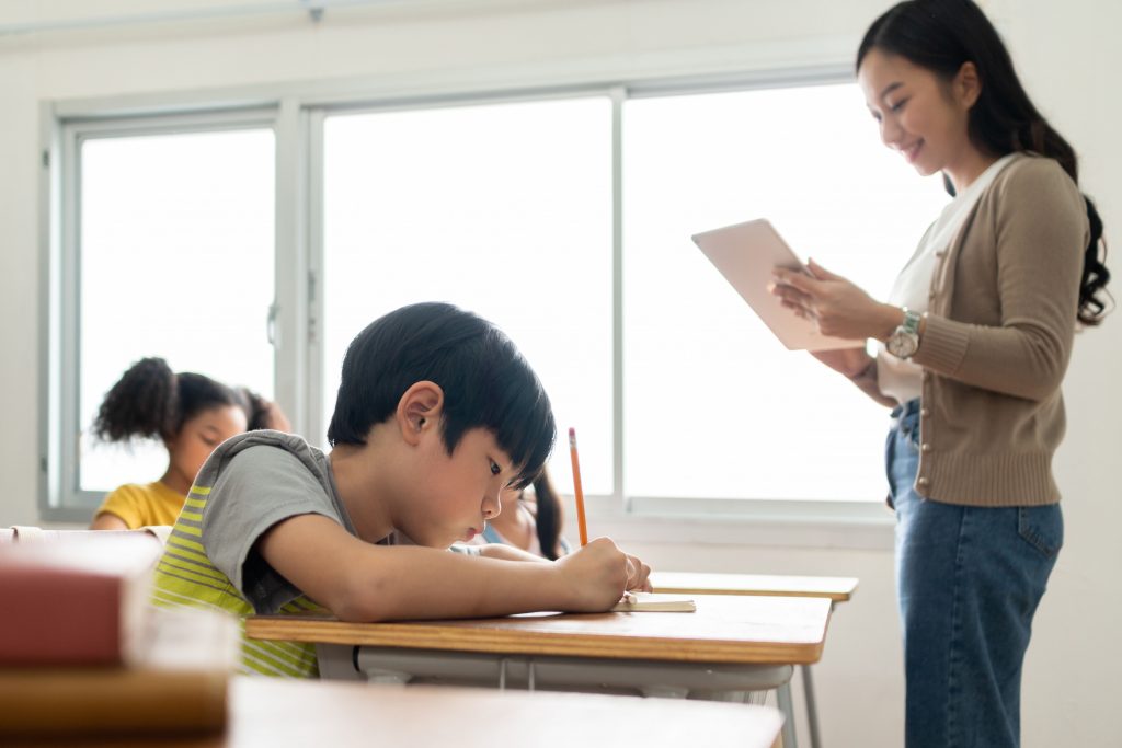 Asian,Children,And,Female,Teacher,In,Classroom,At,Elementary,School. ครูมักให้คะแนนเด็กผู้หญิงมากกว่าเด็กผู้ชาย