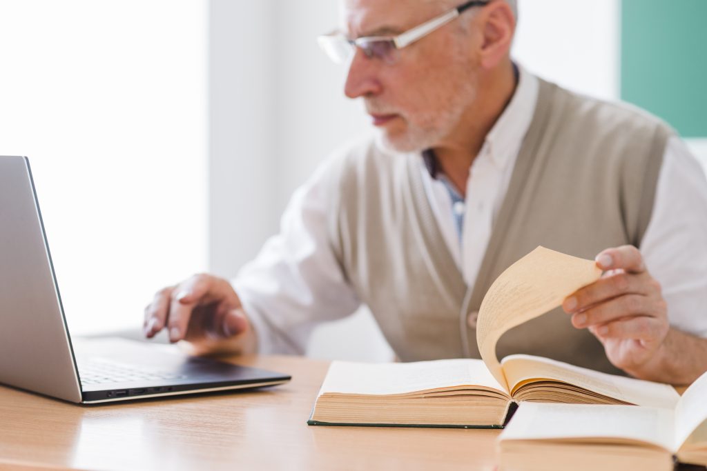 senior-professor-working-with-laptop-while-holding-page-book Live Long Learning
