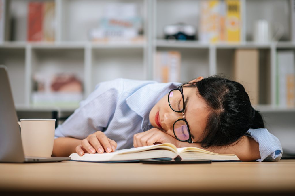 Young woman in eyeglasses sleeping at desk at home เพราะอะไรนักเรียนของครูถึงชอบผัดวันประกันพรุ่ง